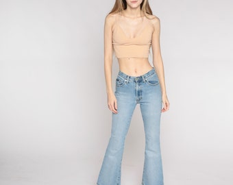May include: A light tan crop top with spaghetti straps and light blue flared jeans. The jeans have a classic five-pocket design. The model is wearing white platform sneakers. The background is plain white.