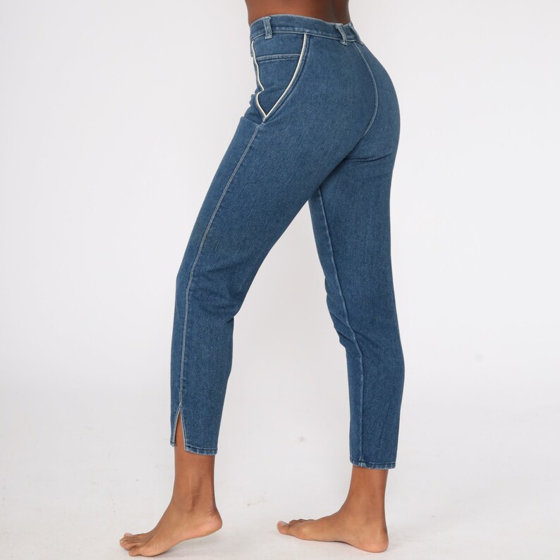 Puede incluir: Pantalones vaqueros azules con ribete blanco a lo largo de los bolsillos y las costuras. Los pantalones tienen una cintura alta y una pernera recta que se estrecha hasta el tobillo, con una peque&ntilde;a abertura en el dobladillo. Fondo blanco.