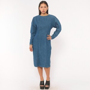 Puede incluir: Un vestido de mezclilla azul de tono medio con mangas largas de hombros ligeramente abullonados, cuello redondo y un corpi&ntilde;o ajustado que se transforma en una falda recta que llega hasta la rodilla. El vestido tiene costuras verticales.