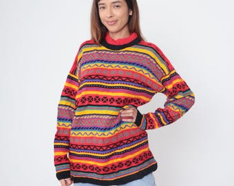 May include: A colorful, patterned sweater with a red base and horizontal stripes in yellow, purple, blue, and black. The sweater has a mock neck and long sleeves with black cuffs. The design includes geometric shapes.