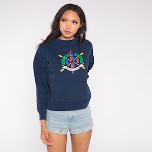 Puede incluir: Sudadera azul marino con cuello redondo y un gráfico colorido en la parte delantera. El gráfico incluye remos cruzados, una corona de hojas y el texto "HOOK SPORT". La sudadera se combina con pantalones cortos vaqueros de color azul claro.