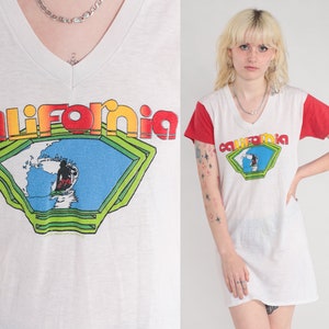 K&ouml;nnte beinhalten: Wei&szlig;es T-Shirt mit einem rot-wei&szlig;en Farbblock-Design. Das Shirt hat einen V-Ausschnitt und zeigt ein Grafikbild eines Surfers, der auf einer Welle reitet, mit dem Wort "California" in einer Regenbogen-Schrift.