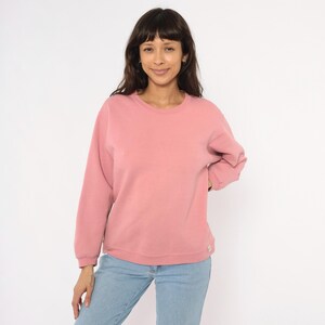 May include: A solid rose-colored sweatshirt with a crew neck and long sleeves. The sweatshirt is paired with light blue jeans. The model is smiling and looking at the camera.