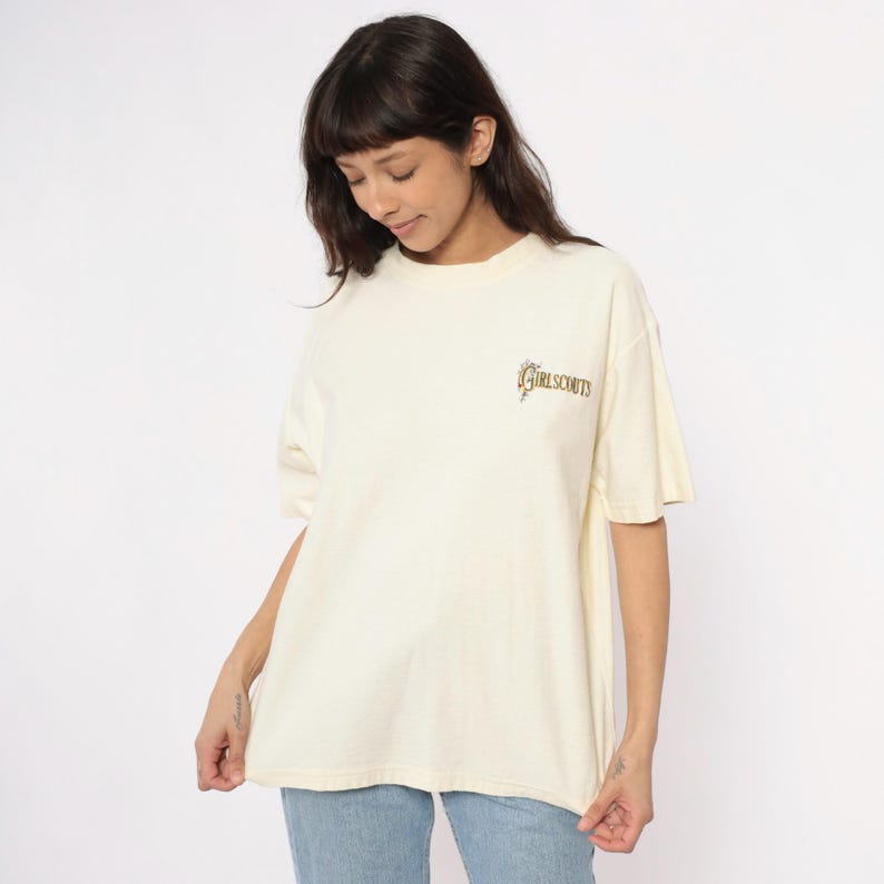 May include: Cream-colored t-shirt with short sleeves. The shirt features the text "GIRL SCOUTS" in a brown, stylized font on the upper chest. The model is wearing blue jeans. The shirt is a casual, everyday wear item.