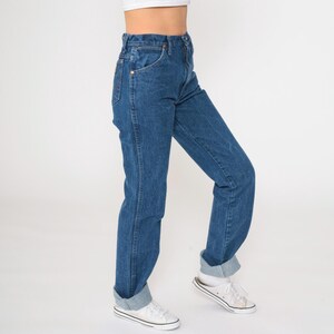 May include: A pair of blue denim jeans with a high waist and a straight leg fit. The jeans are cuffed at the bottom and the model is wearing white sneakers.