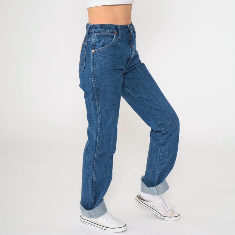 May include: A pair of blue denim jeans with a high waist and a straight leg fit. The jeans are cuffed at the bottom and the model is wearing white sneakers.