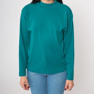 Pode incluir: Camisa de manga comprida verde-azulado com gola alta. A camisa &eacute; feita de um material macio e &eacute; combinada com jeans azuis. A parte superior &eacute; de cor s&oacute;lida e tem um design simples e cl&aacute;ssico.
