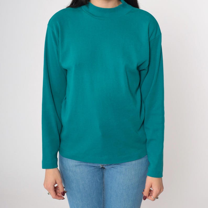 Pode incluir: Camisa de manga comprida verde-azulado com gola alta. A camisa &eacute; feita de um material macio e &eacute; combinada com jeans azuis. A parte superior &eacute; de cor s&oacute;lida e tem um design simples e cl&aacute;ssico.