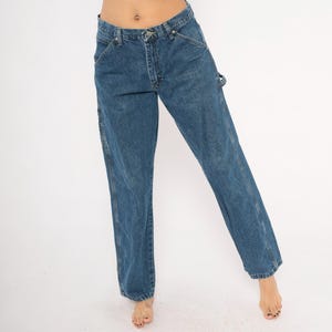 May include: Blue denim carpenter jeans with a straight leg fit. The jeans have classic five-pocket styling, a button and zipper closure, and a hammer loop. The denim is a medium wash with subtle fading and wear.