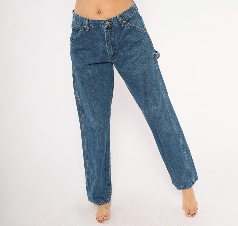 May include: Blue denim carpenter jeans with a straight leg fit. The jeans have classic five-pocket styling, a button and zipper closure, and a hammer loop. The denim is a medium wash with subtle fading and wear.