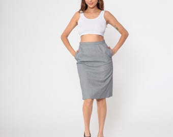 May include: A woman wearing a white tank top and a gray pencil skirt with pockets. She is also wearing black platform heels. The outfit is set against a white background.