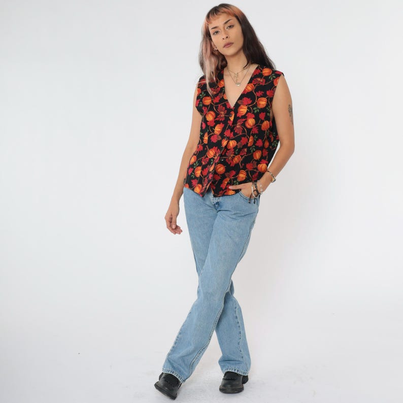 Puede incluir: Una blusa negra sin mangas con estampado floral rojo y naranja. La blusa tiene un escote en V y se lleva con jeans azul claro.