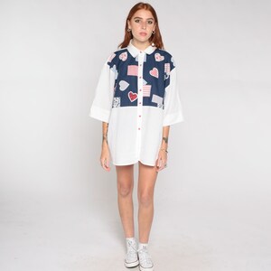 May include: A white button-down shirt with a blue and red patchwork design featuring hearts and squares. The shirt has a short sleeve and is worn by a person with red hair.
