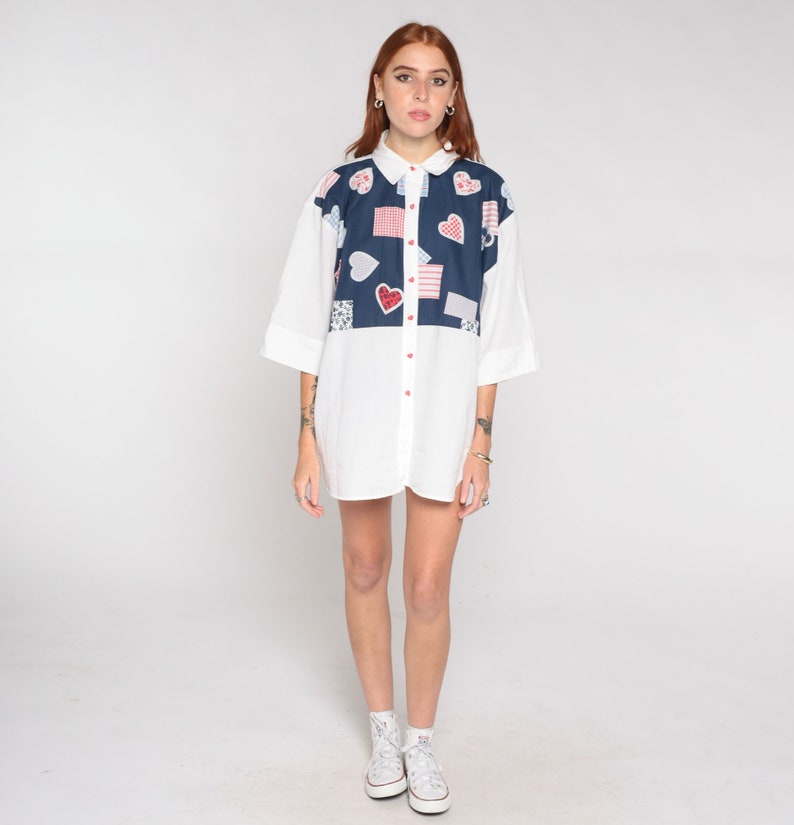 May include: A white button-down shirt with a blue and red patchwork design featuring hearts and squares. The shirt has a short sleeve and is worn by a person with red hair.