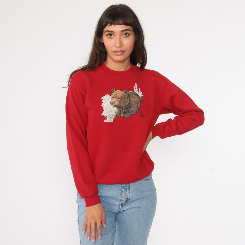May include: Red sweatshirt with a brown bear graphic on the front. The bear is surrounded by white and brown foliage. The sweatshirt has a crew neck and long sleeves.