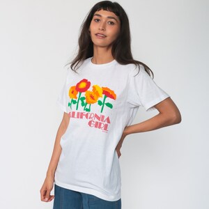 Könnte beinhalten: Weißes T-Shirt mit einem Aufdruck von bunten Blumen und dem Text "CALIFORNIA GIRL".