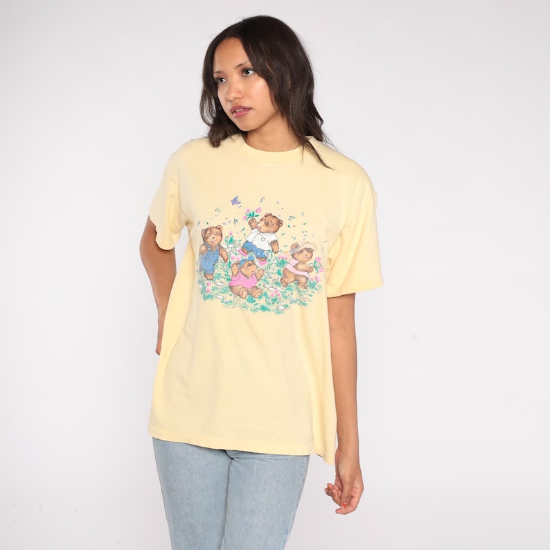 May include: A light yellow t-shirt featuring a graphic of four teddy bears playing in a field of flowers. The bears are wearing different outfits. The shirt is a casual, short-sleeved style.