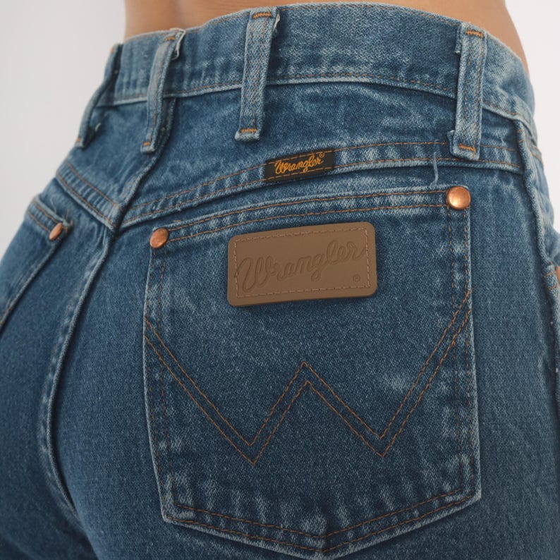 May include: Close-up of blue denim jeans with the Wrangler logo on the back pocket. The jeans have copper-colored rivets and a brown leather patch with the Wrangler brand name. The stitching is a contrasting brown color.
