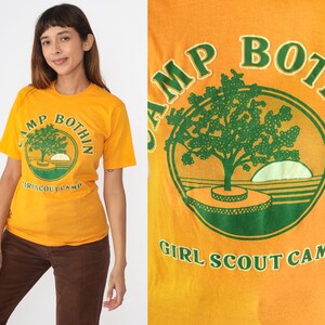 May include: Mustard yellow t-shirt with a green and orange graphic. The graphic features a tree, a setting sun, and the text "CAMP BOTHIN GIRL SCOUT CAMP" in a circular design. The shirt has short sleeves.