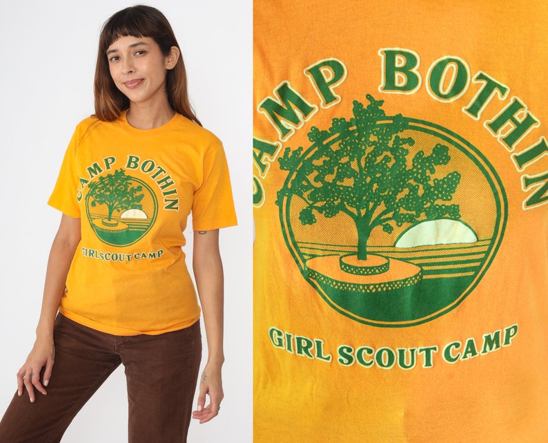 May include: Mustard yellow t-shirt with a green and orange graphic. The graphic features a tree, a setting sun, and the text "CAMP BOTHIN GIRL SCOUT CAMP" in a circular design. The shirt has short sleeves.