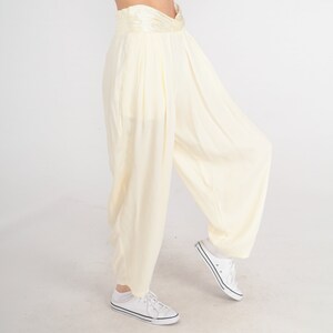 May include: Cream-colored, wide-leg pants with a high, gathered waistband. The pants have a loose, flowing design and are paired with white sneakers. The fabric appears to be lightweight and suitable for warm weather.