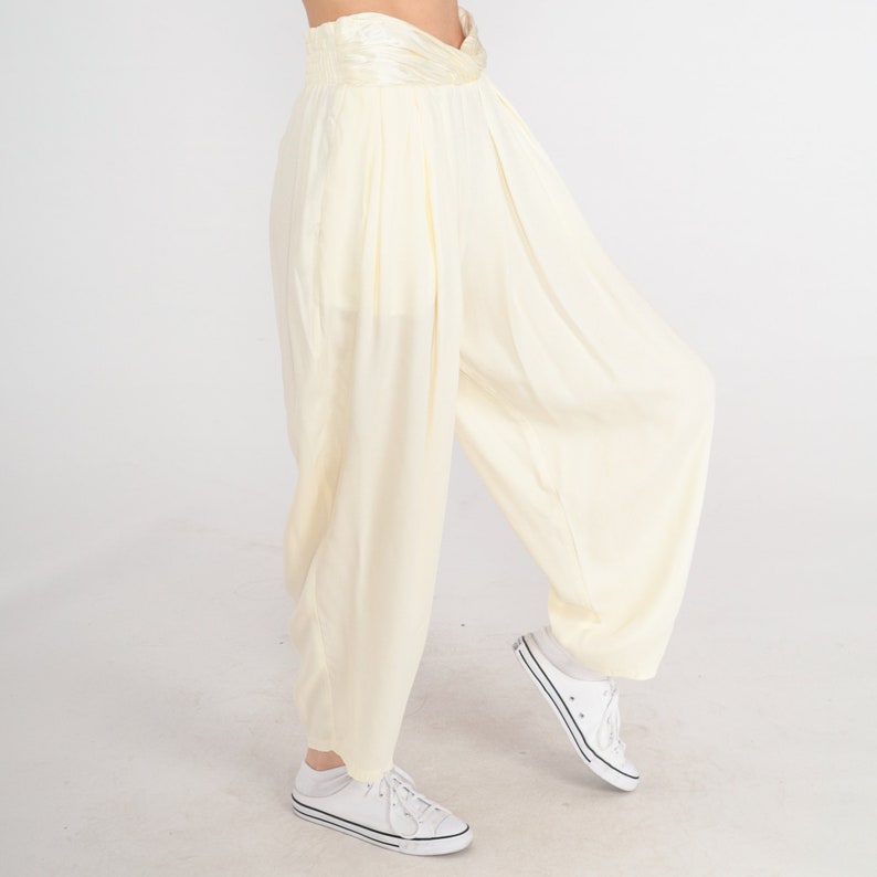 May include: Cream-colored, wide-leg pants with a high, gathered waistband. The pants have a loose, flowing design and are paired with white sneakers. The fabric appears to be lightweight and suitable for warm weather.