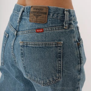 Peut inclure: Jean en denim bleu clair avec une conception classique &agrave; cinq poches. Le dos pr&eacute;sente un patch en cuir marron avec le logo "Wrangler" et une petite &eacute;tiquette rouge "Wrangler" sur la poche arri&egrave;re. Le jean a une coupe droite.