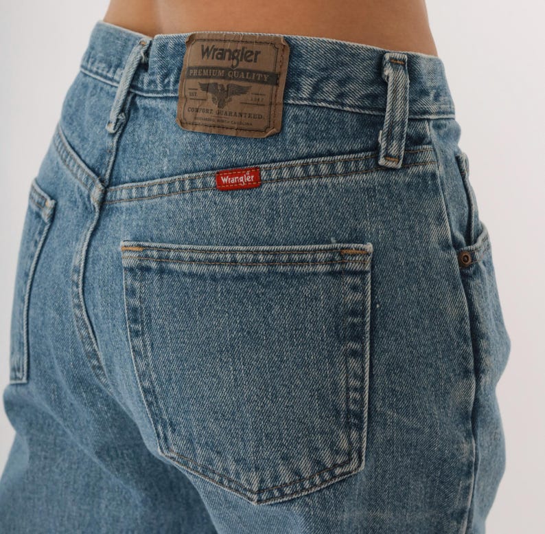 Peut inclure: Jean en denim bleu clair avec une conception classique &agrave; cinq poches. Le dos pr&eacute;sente un patch en cuir marron avec le logo "Wrangler" et une petite &eacute;tiquette rouge "Wrangler" sur la poche arri&egrave;re. Le jean a une coupe droite.