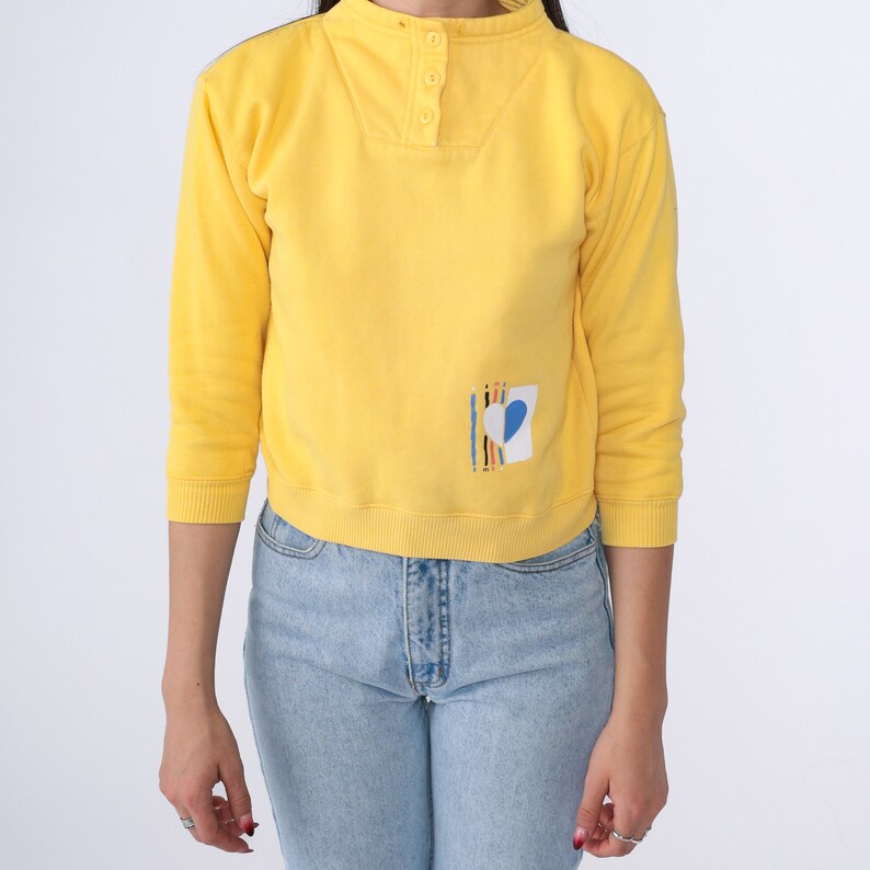 May include: A yellow cropped sweatshirt with a blue heart and colourful stripes on the right side. The sweatshirt has a mock neck and a button closure at the neck.