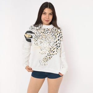 May include: A white long-sleeve sweatshirt featuring a large graphic of a leopard face and spots in black and gold tones. The design extends across the front and onto the right sleeve, incorporating a metallic gold accent near the shoulder area.