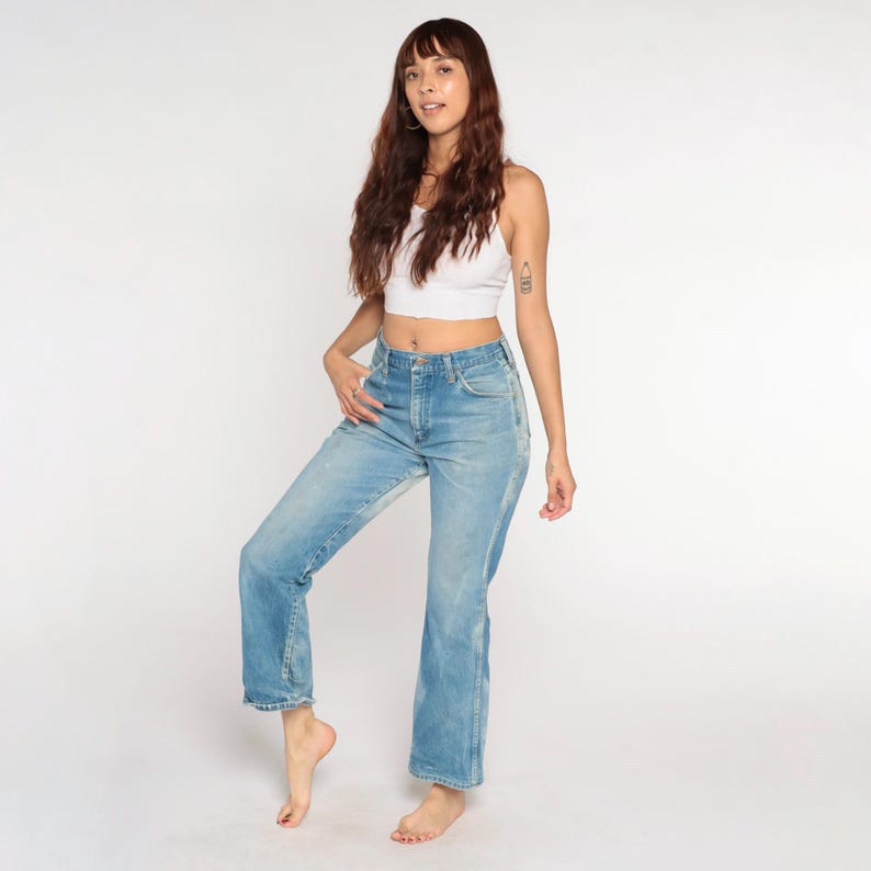 May include: Light blue denim jeans with a high waist and flared leg. The jeans have a classic five-pocket design and a faded wash. The model is wearing a white vest top.