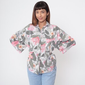 Könnte beinhalten: Ein Hemd mit Knopfleiste und floralem Muster in Rosa, Grau und Weiß. Das Hemd hat eine lockere Passform mit weiten Ärmeln. Das florale Design umfasst große rosa Blüten und graue Blätter. Das Hemd wird mit hellblauen Jeans getragen.