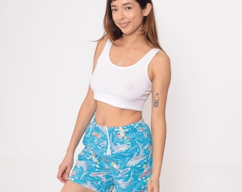 May include: A woman wearing a white crop top and blue swim shorts with a colorful fish and dolphin print.