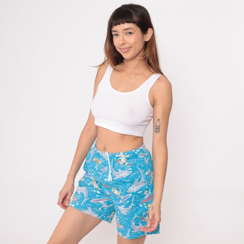 K&ouml;nnte beinhalten: Eine Frau tr&auml;gt ein wei&szlig;es Crop-Top und blaue Badeshorts mit einem bunten Fisch- und Delfin-Druck.