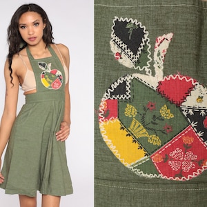 May include: Olive green apron dress with a patchwork apple design on the front. The apple is made of various fabric patches in red, yellow, black, and green, with white stitching. The dress has a halter neckline and a flared skirt.