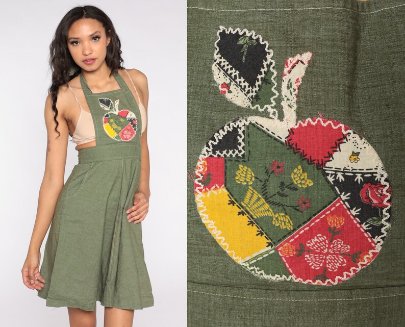 May include: Olive green apron dress with a patchwork apple design on the front. The apple is made of various fabric patches in red, yellow, black, and green, with white stitching. The dress has a halter neckline and a flared skirt.