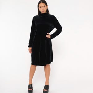 Könnte beinhalten: Ein schwarzes Samtkleid mit hohem Ausschnitt und langen Ärmeln. Das Kleid reicht bis zu den Knien und wird mit schwarzen Plateau-Heels getragen. Das Model posiert vor einem weißen Hintergrund.