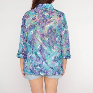 Op de afbeelding: Een doorschijnende blouse met lange mouwen en een levendig, abstract patroon in tinten blauw, paars en blauwgroen. De blouse heeft een kraag en driekwart mouwen. Het kledingstuk wordt gedragen met een spijkerbroekshort.