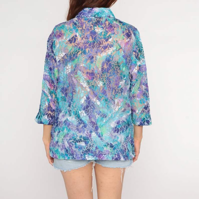 Op de afbeelding: Een doorschijnende blouse met lange mouwen en een levendig, abstract patroon in tinten blauw, paars en blauwgroen. De blouse heeft een kraag en driekwart mouwen. Het kledingstuk wordt gedragen met een spijkerbroekshort.