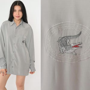 May include: A gray long-sleeved button-down shirt with a crocodile logo embroidered on the front. The logo is surrounded by a decorative border.
