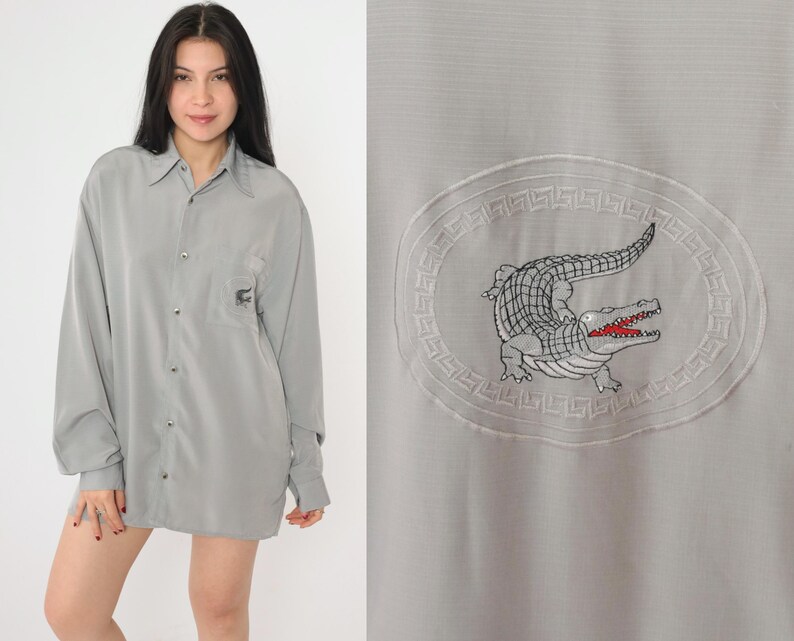 May include: A gray long-sleeved button-down shirt with a crocodile logo embroidered on the front. The logo is surrounded by a decorative border.