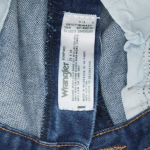 May include: A blue denim Wrangler jeans tag with white text. The tag says "Machine wash cold inside out with like colors. Only non-chlorine bleach when needed, tumble dry low or line dry. Made in Nicaragua of US fabric. 100% cotton. Exclusive of decoration."