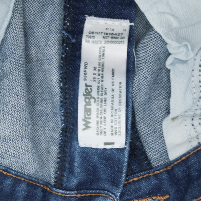 May include: A blue denim Wrangler jeans tag with white text. The tag says "Machine wash cold inside out with like colors. Only non-chlorine bleach when needed, tumble dry low or line dry. Made in Nicaragua of US fabric. 100% cotton. Exclusive of decoration."