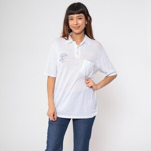 May include: White polo shirt with a pocket and short sleeves. The shirt has a blue graphic on the chest. The collar and pocket are the same color as the shirt. The shirt is worn with blue jeans.