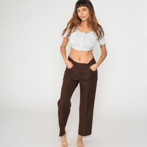 May include: A light gray, short-sleeved crop top with a button-up front and brown pants. The pants are a solid color and have a straight leg design. The outfit is simple and casual.