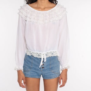 May include: White long-sleeved blouse with a ruffled neckline and lace trim. The blouse has a drawstring at the waist and is worn with denim shorts.
