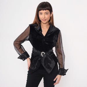 May include: Black velvet blouse with sheer sleeves and a belted waist. The blouse features a V-neckline and a decorative buckle on the belt. The sleeves have buttoned cuffs. The blouse is paired with black pants.
