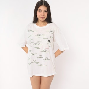 May include: A white short-sleeved t-shirt featuring a repeating pattern of small black-outlined sheep grazing on green grass patches. The word "Solomon" is printed in red script on the front. The shirt has a crew neckline and a loose fit.