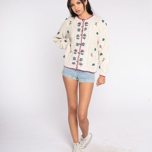 May include: A white knitted cardigan with a cable knit pattern and floral embroidery. The cardigan has a pink trim and buttons. The model is wearing denim shorts and white sneakers.