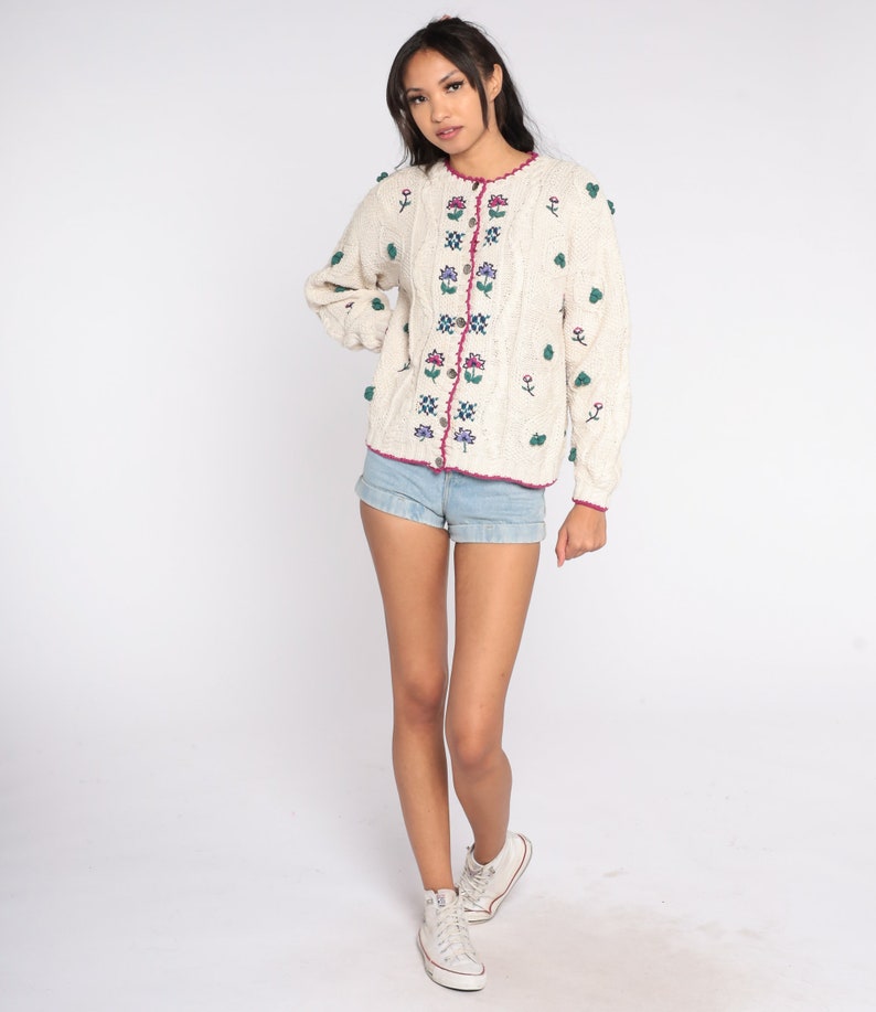 May include: A white knitted cardigan with a cable knit pattern and floral embroidery. The cardigan has a pink trim and buttons. The model is wearing denim shorts and white sneakers.